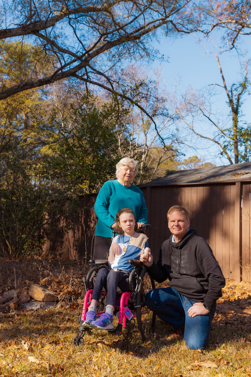 A young girl sits in a wheelchair outside with her grandmother and father surrounding her. 