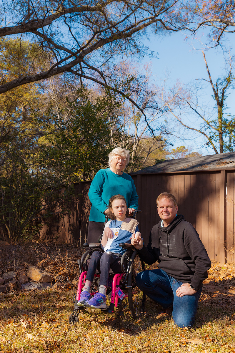 A young girl sits in a wheelchair outside with her grandmother and father surrounding her. 