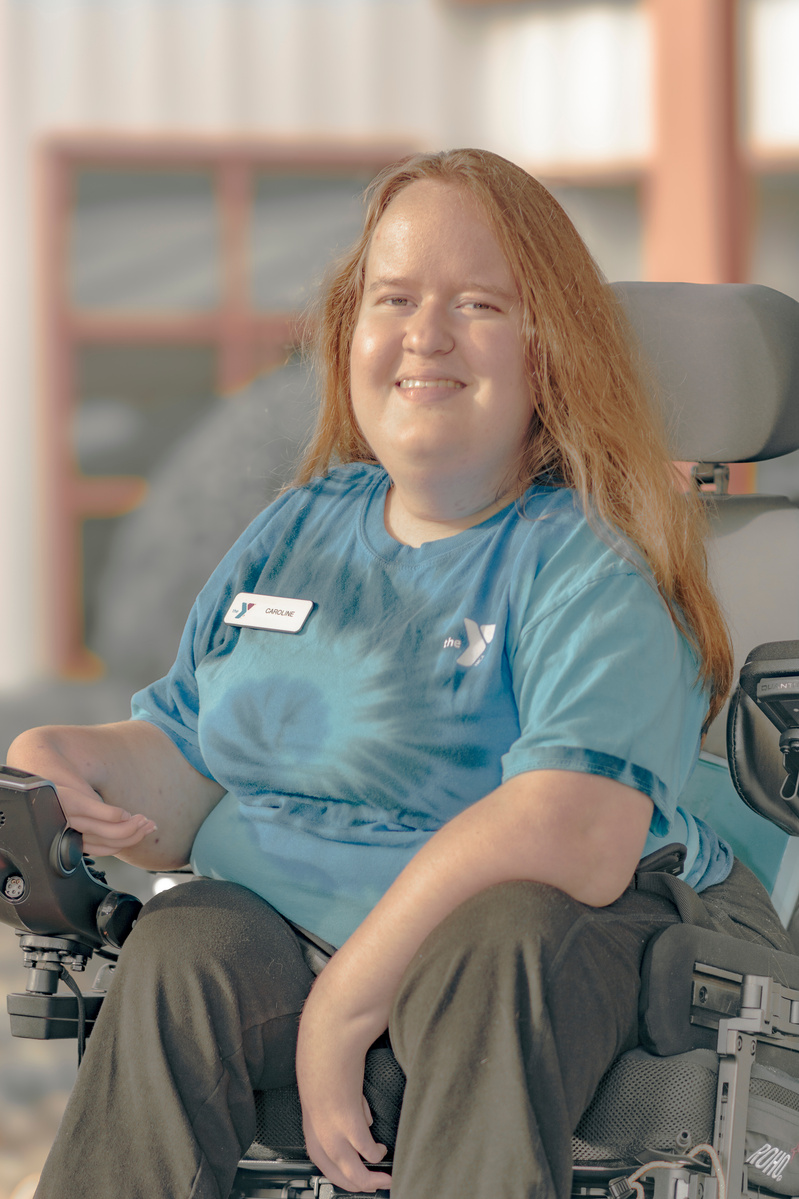 Woman with red hair wears a blue t-shirt and a name tag that reads Caroline. She is sitting in a power wheelchair. 
