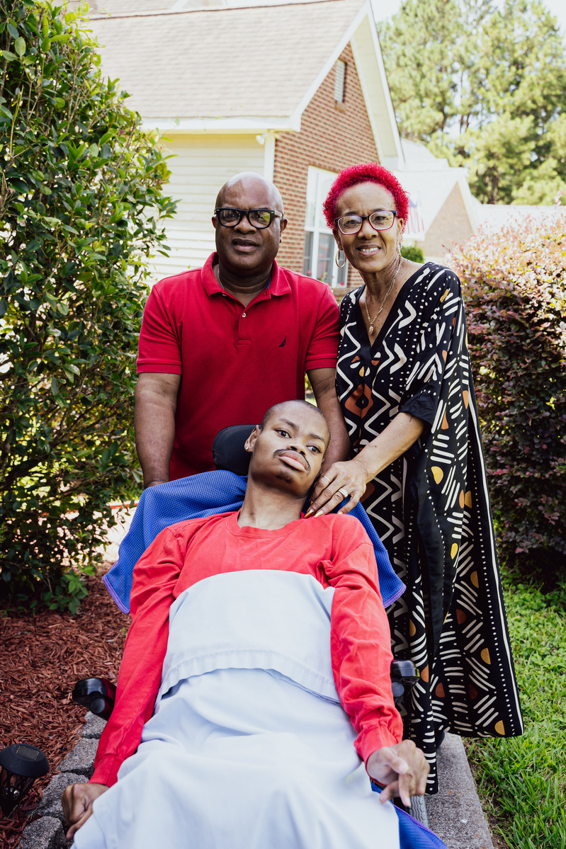 A mother and father stand behind their son as he sits in a wheelchair. 