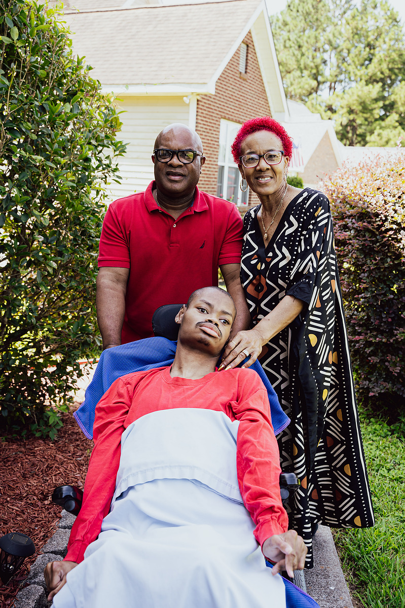 A mother and father stand behind their son as he sits in a wheelchair. 