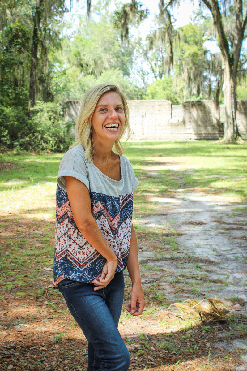 A blond woman in a patterned blouse and jeans stand outside and smiles for the camera. 