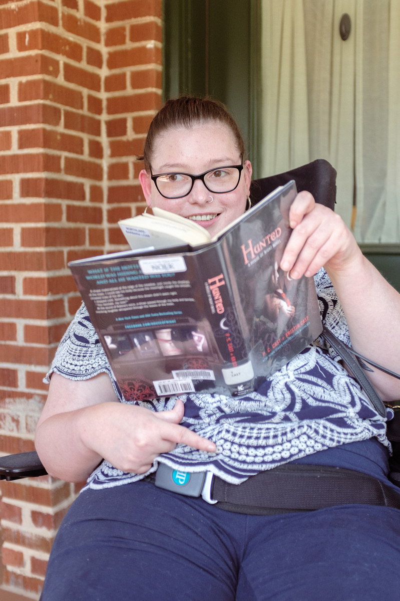 A young brunette woman in a patterned shirt and glasses holds a book and sits on a screened porch. 