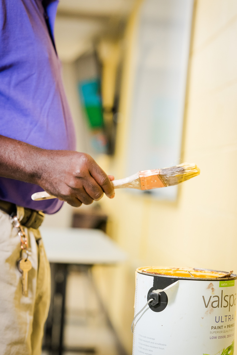 A middle aged African American man wears a purple polo shirt and paints a wall cream.