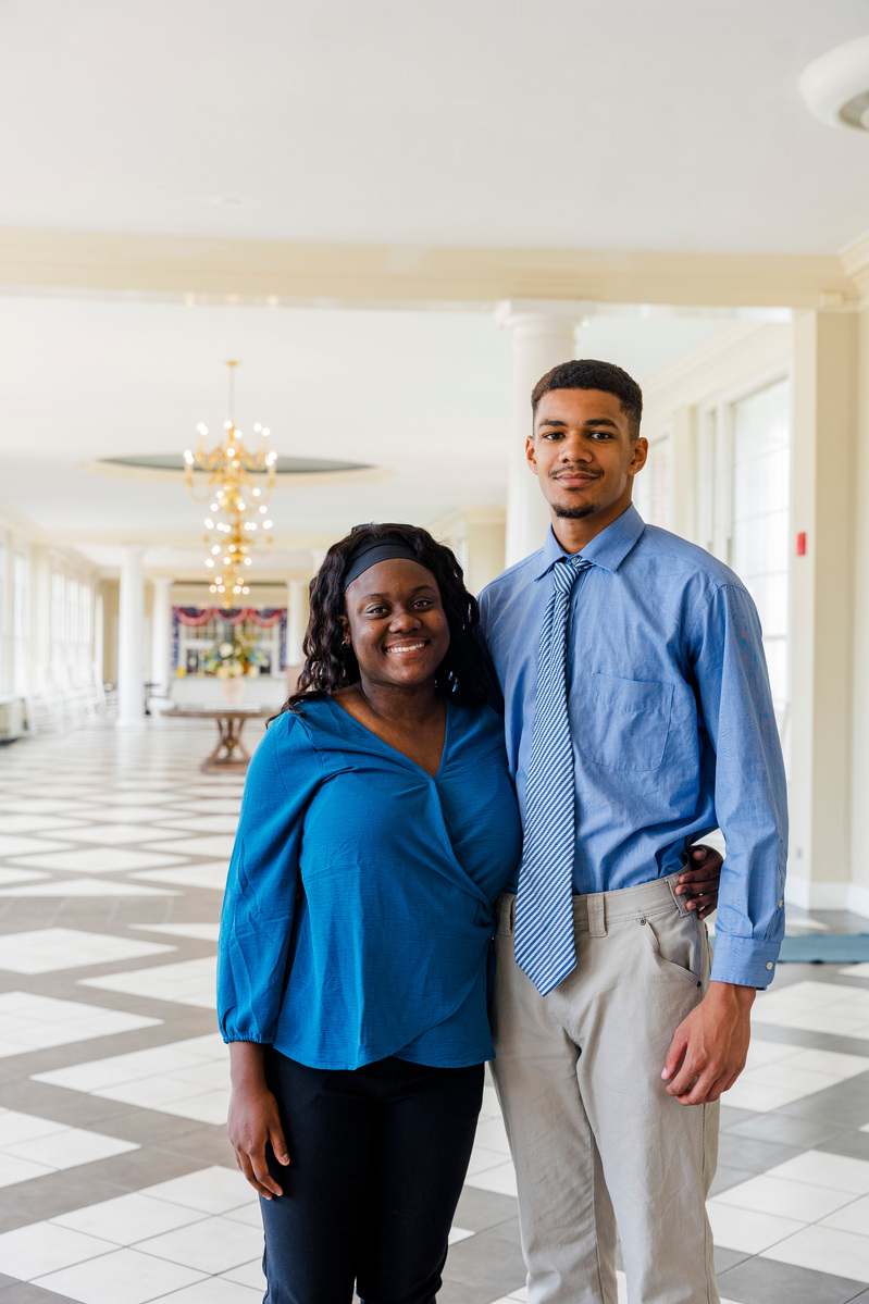 Two students wear blue shirts and stand in a bright hallway. 