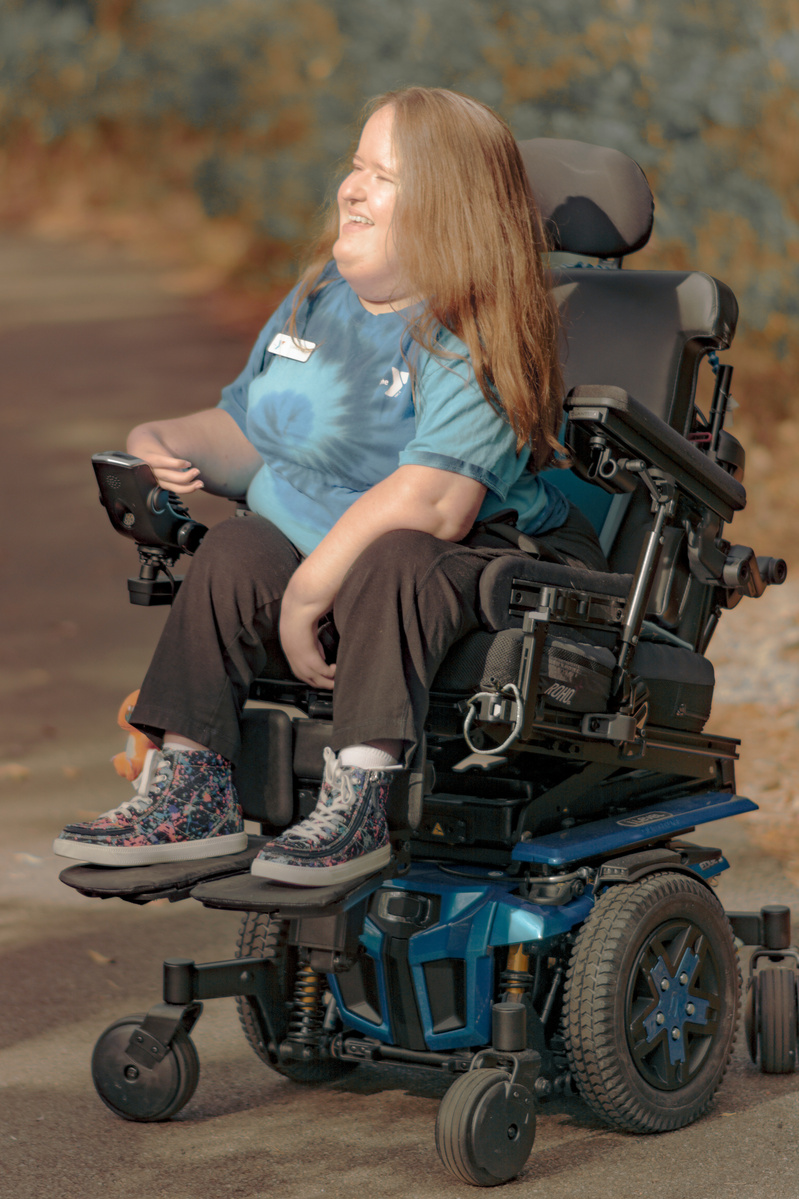 Woman with red hair wears a blue t-shirt is sits in her power wheelchair and smiles.  