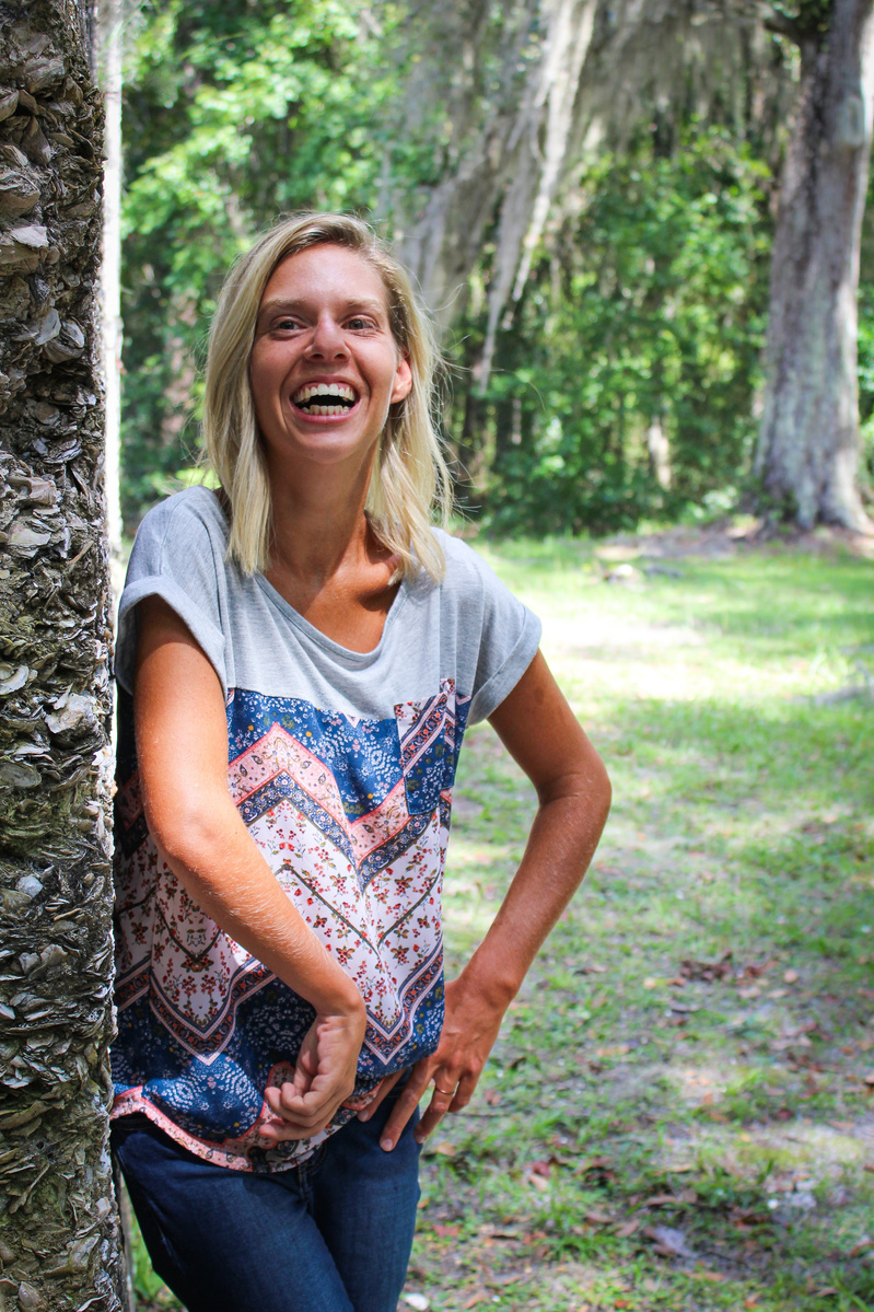 A blond woman in a patterned blouse and jeans stand outside and smiles for the camera. 