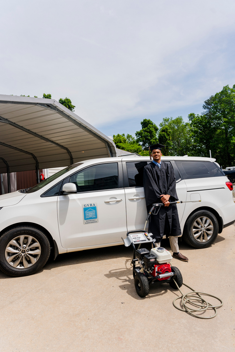 A young African American male wears a cap and gown as he stands in front of a white van and holds a pressure washier. 