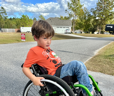 A young boy sits outside in an orange shirt and leans back in his wheelchair 