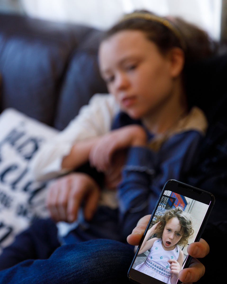 A phone shows a photo of a toddler making a goofy face. The same girl, now 11, sits behind the phone. 