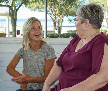 Two women with blond hair sit outside and talk to each other.
