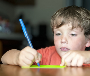 A young boy wears an orange shirt and sits at a table writing.  
