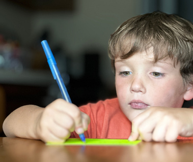 A young boy wears an orange shirt and sits at a table writing.  