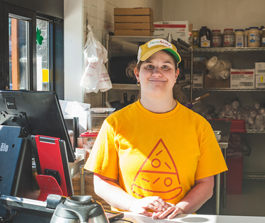 A young woman wears a yellow shirt with a drawing of a pizza slice on the front of it. She works at a restaurant register.