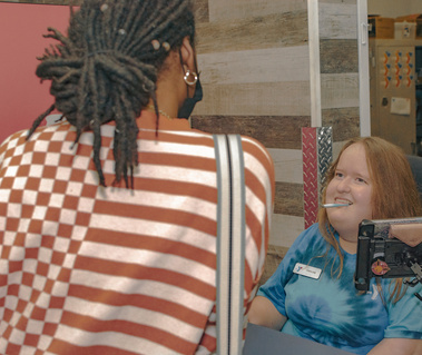 Woman with red hair wears a blue t-shirt and speaks to another woman who has her back to the camera. 