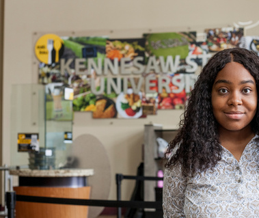 A young African American woman named Skye wears a white button front blouse and stands in front of a Kennesaw State University sign. 