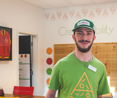 Young man wears green t-shirt and a baseball cap as he stands in front of a work board. 