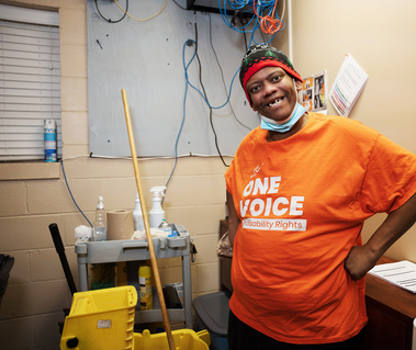 A man in an orange shirt and knit cap has his hands on his hip and smiles for the camera. 