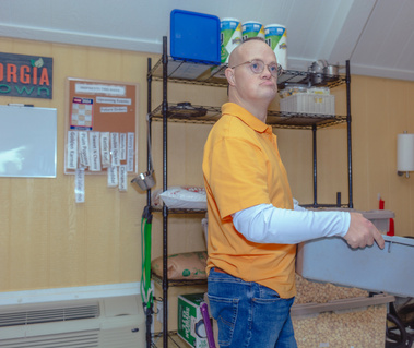Man wears yellow shirt and jeans and carries a grey plastic bin. 