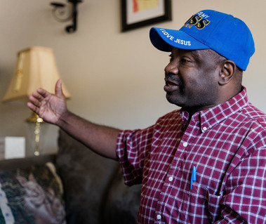 African American man in a plaid shirt and blue ball cap smiles in his living room.