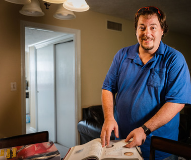 A man stands in a blue polo shirt and looks at a book on a table. 