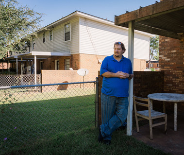 A man in a blue polo shirt leans against a patio post and looks out across his yard. 