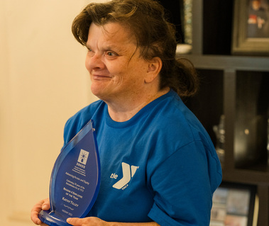 A brunette woman wears a blue t-shirt and holds a glass award. 