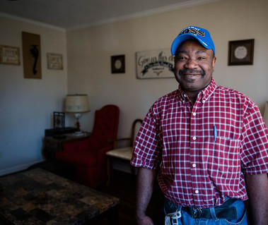 African American man in a plaid shirt and blue ball cap smiles in his living room.