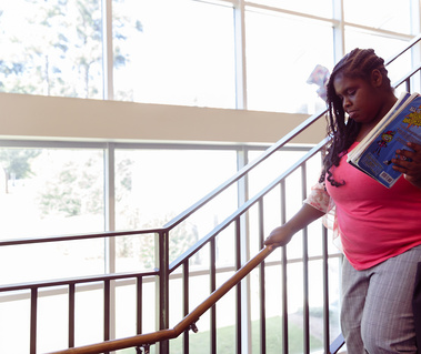 An African American woman wears a pink shirt and grey pants as she walks down a flight of stairs holding a stack of books. 