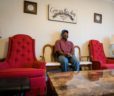 African American man in a plaid shirt and blue ball cap smiles in his living room.