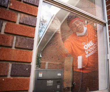 A man in an orange shirt and a knit cap cleans a window and looks outside. 