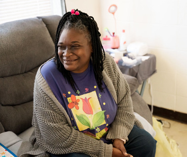 A black woman with braids and a purple shirt looks to her right as she sits on a sofa.