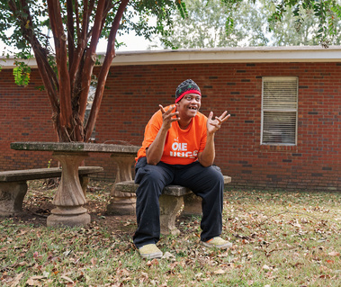 A man in an orange shirt and knit cap sits outside on a bench smiling.