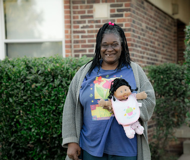 A black woman with braids and a purple shirt holds a doll as she stands outside and smiles. 
