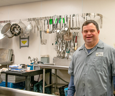 A man wearing a grey shirt stands in a kitchen in front of a sink and utensils.