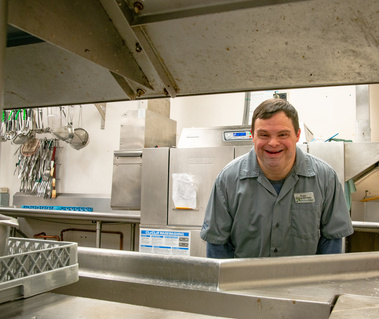 A man in a grey collared shirt stands in a kitchen and looks across a counter while smiling. 