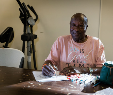 A man sits at a table in a pink shirt drawing pictures. 