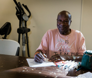 A man sits at a table in a pink shirt drawing pictures. 