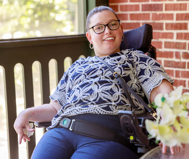 A young brunette woman in a patterned shirt and glasses sits in a wheelchair next to flowers. 