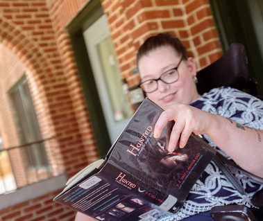 Young brunette woman in a patterned shirt and glasses sits on a screened porch while reading a book.
