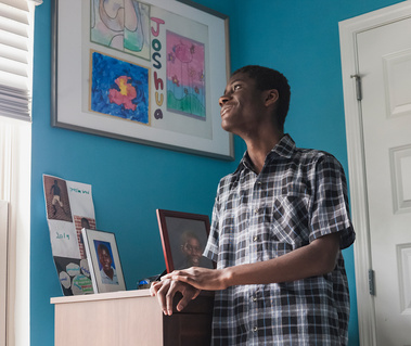 A young African American man wears a plaid shirt and smiles while standing in his bedroom.
