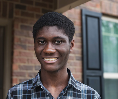 A young African American man wears a plaid shirt and smiles in front of his home.