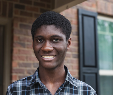 A young African American man wears a plaid shirt and smiles in front of his home.