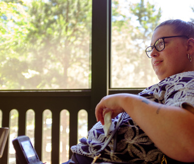 Young brunette woman in a patterned shirt and glasses sits on a screened porch. 