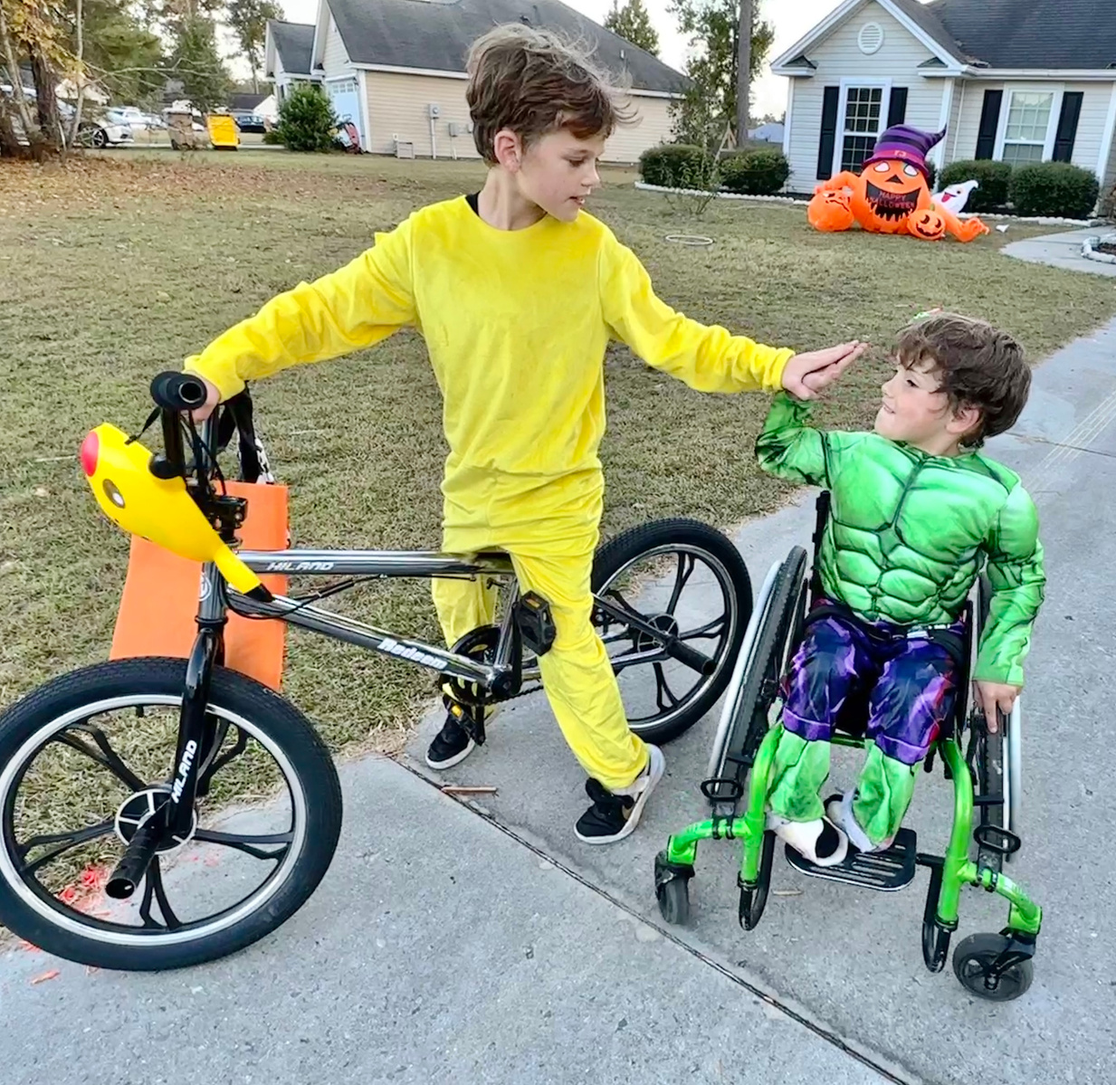 Tow boys wear Halloween costumes and high five each other on the sidewalk. 