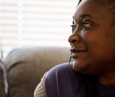 A black woman with braids and a purple shirt looks to her right as she sits on a sofa.