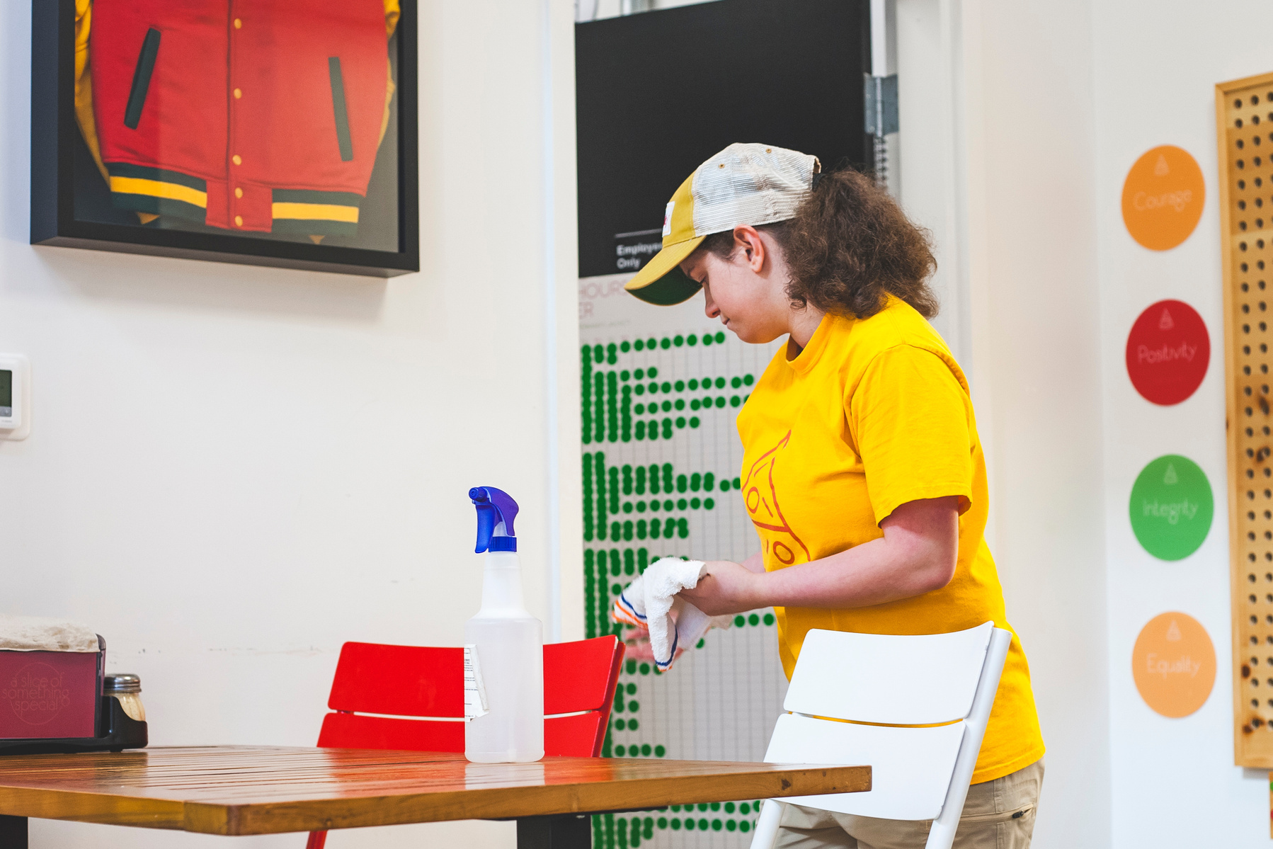 A young woman in a yellow hat and t-shirt clears a restaurant table. 