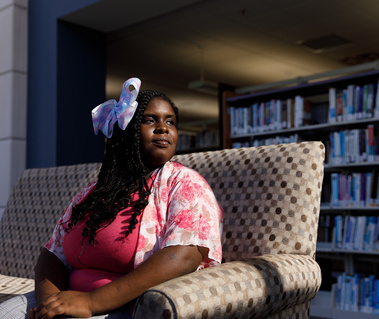 An African American woman with bow in her hair sits on a sofa in front of shelves filled with books.