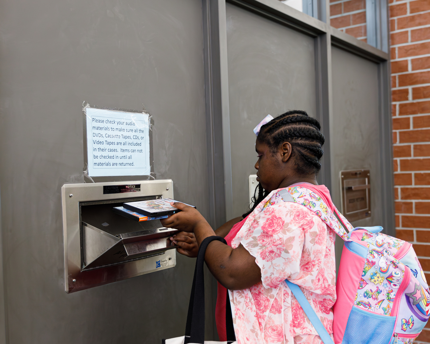A young African American woman with braids in her hair and a pink flowered shirt puts books into a library return bin.