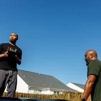 A young man stands on a trampoline in front of a blue sky while his father stands near him. 